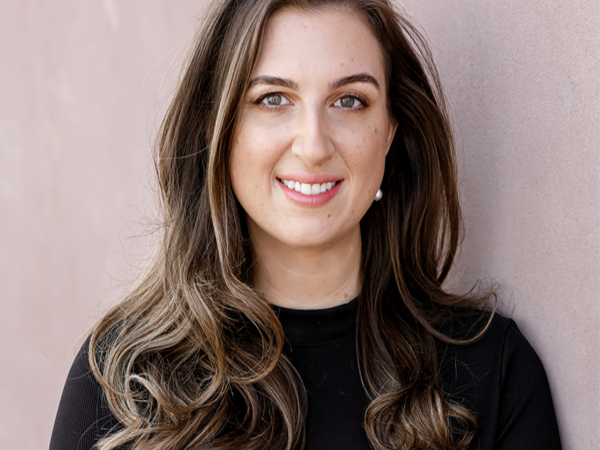 Female with brown hair in black top poses in front of pink wall.